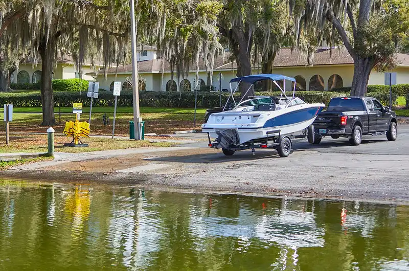 Black pickup truck backing a white and blue motorboat on a trailer down a boat ramp toward the water at a lakeside marina surrounded by trees and buildings.